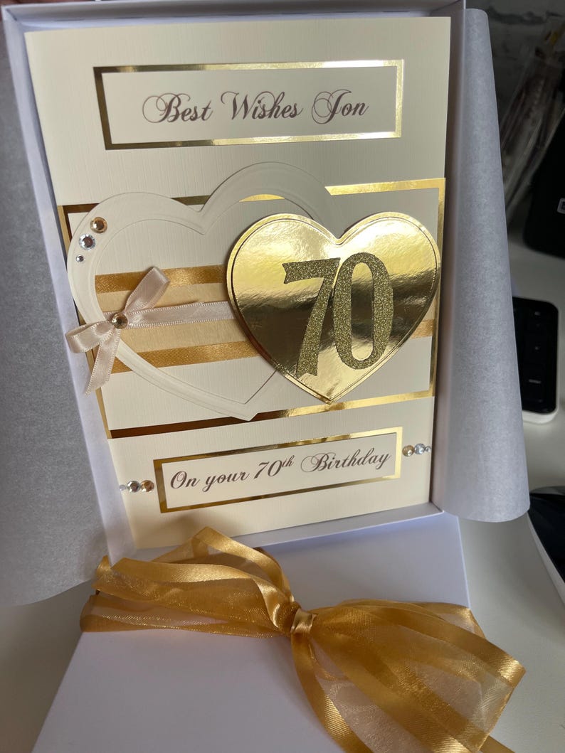 May include: A white card with gold accents and a gold heart with the number 70 on it. The card says "Best Wishes Son" and "On your 70th Birthday".