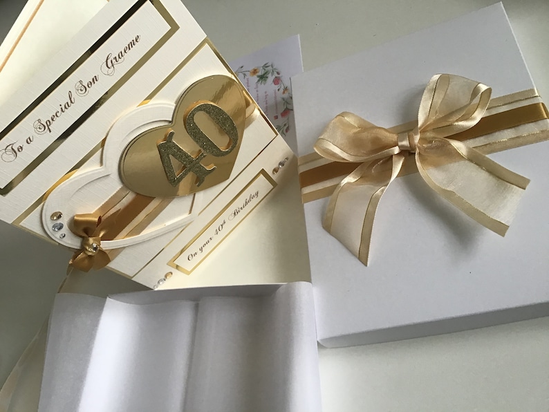 May include: A white card with gold accents and a gold heart with the number 40 on it. The card says "To a special Son Graeme" and "On your 40th Birthday". The card is in a white box with a gold ribbon.
