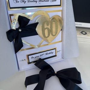 May include: A white card with a black and gold border. The card has a gold heart with the number 60 on it and a black bow. The text on the card reads "To My Darling Husband Alan On your 60th Birthday".