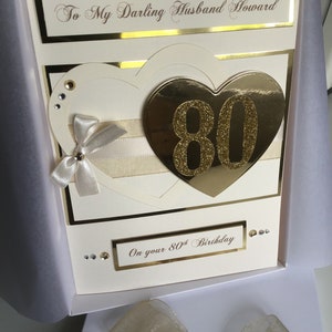 May include: A white card with gold accents and a gold heart with the number 80 on it. The card says "To My Darling Husband Howard" and "On your 80th Birthday".