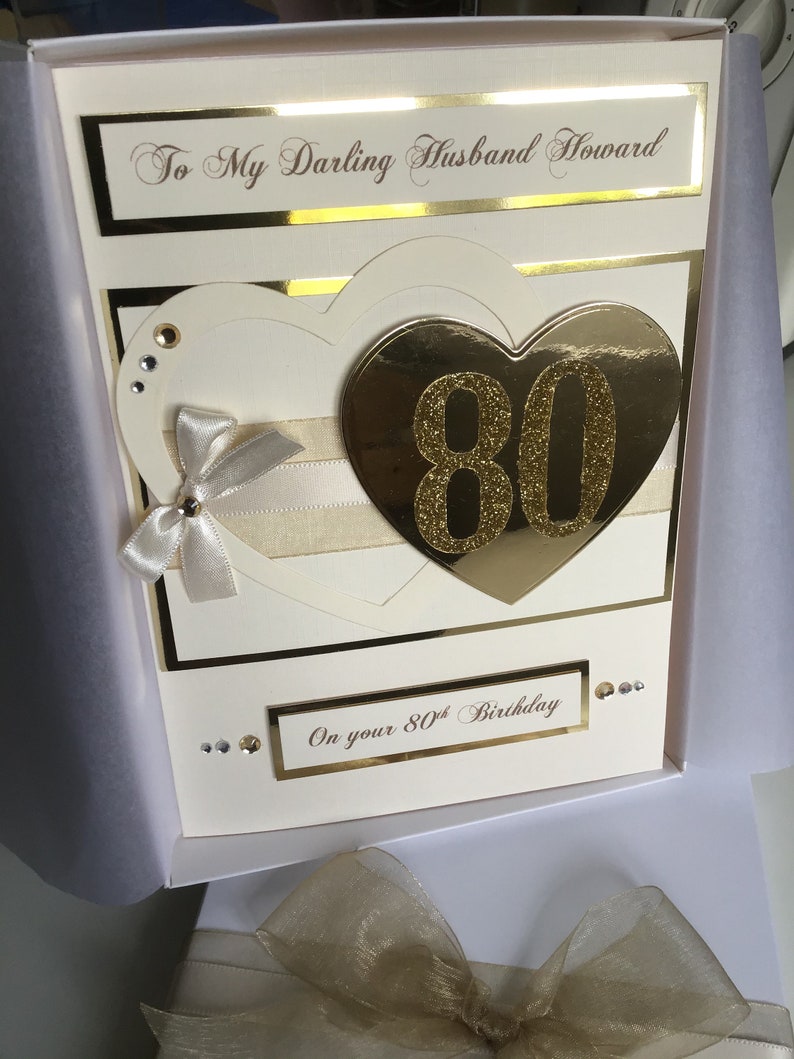 May include: A white card with gold accents and a gold heart with the number 80 on it. The card says "To My Darling Husband Howard" and "On your 80th Birthday".