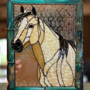 May include: Stained glass panel featuring a horse's head. The artwork uses shades of cream, tan, and black, with a decorative patterned background. The panel is framed in a teal-green border, creating a striking contrast.