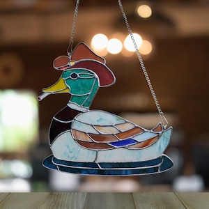 Puede incluir: Pato de vidriera con sombrero de vaquero y fumando un cigarrillo. El pato tiene la cabeza verde, pico amarillo y sombrero marrón. El cuerpo del pato es un mosaico de vidrio azul, marrón y blanco. Cuelga de una cadena plateada.