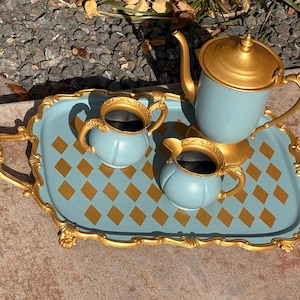 May include: A decorative tea set on a tray. The set includes a light blue teapot and two creamers with gold accents. The tray is light blue with gold diamond patterns and gold handles. The set is designed for serving tea.