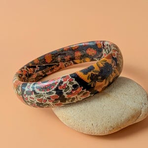 Op de afbeelding: Een ronde armband met een dicht, veelkleurig bloemenpatroon in tinten oranje, zwart, geel en wit. De armband heeft een gladde, glanzende afwerking en rust op een lichtgekleurde, getextureerde, ovaalvormige steen.