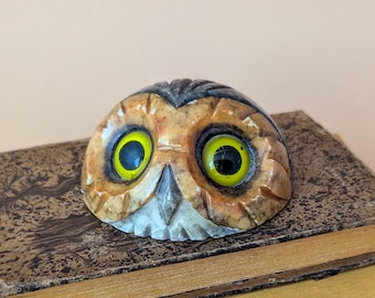 May include: A carved stone owl figurine with prominent yellow eyes and brown and cream mottled markings, resting on top of a stack of two vintage books with textured brown covers and worn yellow edges.