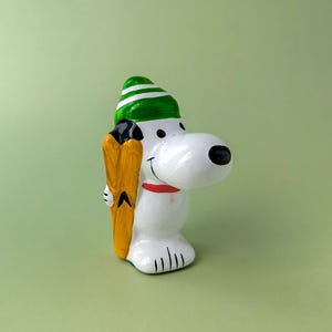 May include: A white ceramic figurine of a cartoon dog wearing a green and white striped hat. The dog has a black nose, a red collar, and is holding a pair of yellow skis against its side. The figure has a smooth, glossy finish and stands upright.
