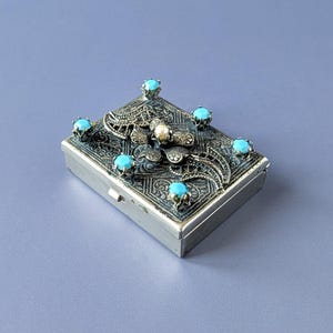 Peut inclure: Une boîte rectangulaire en métal argenté avec un motif en filigrane complexe sur le couvercle. Le couvercle est orné de cinq petites pierres turquoise rondes et vives et d'un accent central blanc nacré sur un motif floral en métal.