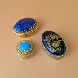 Pode incluir: Três pequenas caixas ovais. Uma é dourada com uma tampa de pedra azul texturizada. Outra é uma caixa dourada menor com um cabochão turquesa brilhante no centro. A terceira é prateada com uma tampa azul escura com detalhes florais dourados.