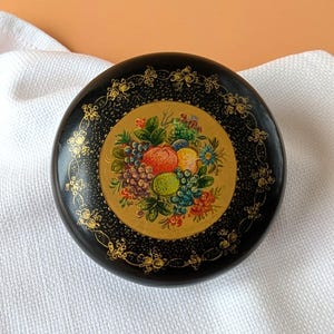 May include: A round black lacquer box featuring a central circular medallion painted with a colourful arrangement of various fruits and green leaves, surrounded by an intricate gold filigree border pattern on the lid's surface.