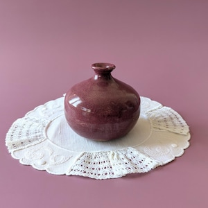 Puede incluir: Un pequeño jarrón de cerámica redondo de color magenta oscuro con acabado brillante y cuello estrecho, apoyado sobre un tapete de tela blanco circular con bordes de encaje intrincados y patrones florales bordados sobre un fondo malva sólido.