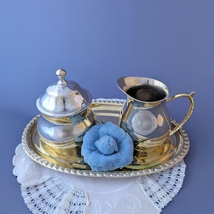 Op de afbeelding: Een verzilverde suikerpot met deksel en een bijpassende melkkan, beide met gepolijste oppervlakken. Deze items zijn gerangschikt op een goudkleurig ovaal dienblad, vergezeld van een decoratieve blauwe stoffen bloem als accent.
