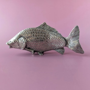 Può includere: Una statuetta di pesce in metallo color argento con dettagliati motivi a scaglie sovrapposte su tutto il corpo, una pinna dorsale prominente e un'ampia coda a ventaglio. La scultura raffigura il pesce in posizione di nuoto.