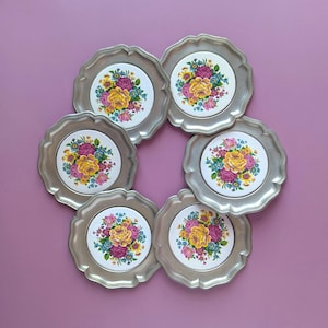 Könnte beinhalten: Ein Set aus sechs runden Metalltellern mit gewelltem Rand, die jeweils eine mittige, weiße Keramikeinlage mit einem lebendigen Blumenstrauß aus gelben, rosa und blauen Blüten in einem klassischen, farbenfrohen botanischen Muster zeigen.