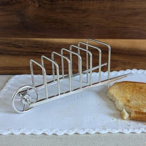 May include: A silver-coloured toast rack shaped like a wheelbarrow. It features multiple slots for holding toast slices. A piece of toasted bread sits beside the rack on a white, lace-edged cloth.