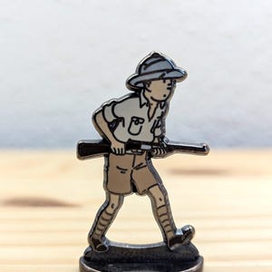 May include: A metal pin featuring a cartoon character in safari attire, holding a rifle. The figure is dressed in a khaki shirt, shorts, and a hat. The pin is silver-toned with black and tan enamel details. The pin is mounted on a small, round base.