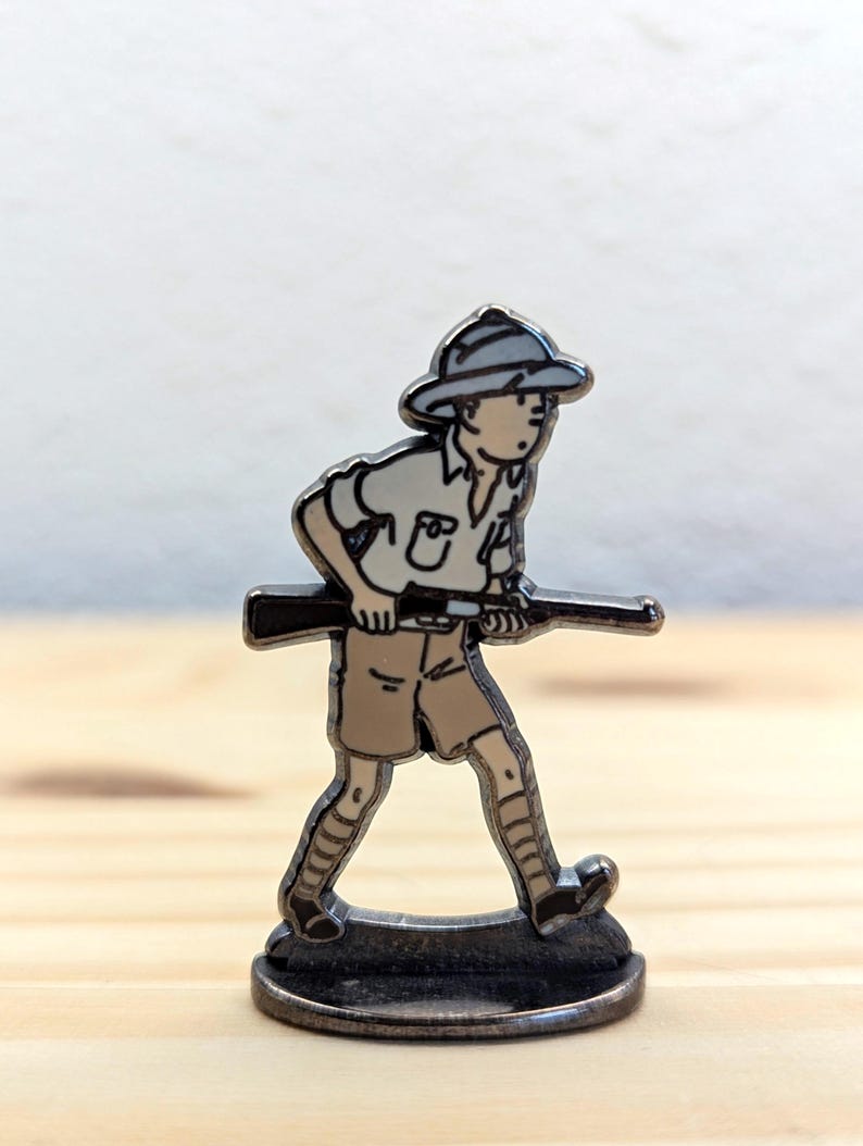 May include: A metal pin featuring a cartoon character in safari attire, holding a rifle. The figure is dressed in a khaki shirt, shorts, and a hat. The pin is silver-toned with black and tan enamel details. The pin is mounted on a small, round base.