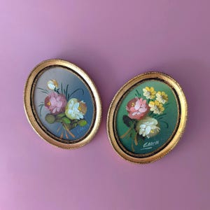 May include: Two oval-shaped wall art pieces featuring hand-painted floral arrangements in gold-toned frames. The left piece has a blue background with pink and white flowers, while the right piece has a green background with pink and yellow flowers.