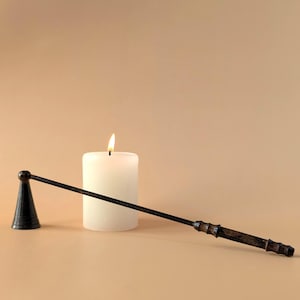 Könnte beinhalten: Ein schwarzer Kerzenlöscher aus Metall mit einem langen, schlanken Griff und einem konischen, glockenförmigen Kopf, der horizontal neben einer weißen Stumpenkerze mit brennender Flamme liegt und eine dunkle, texturierte Oberfläche aufweist.