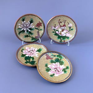 May include: Four round metal plates featuring cloisonné enamel work. Each plate has a yellow background with a repeating geometric pattern and displays colorful floral motifs including pink, white, and purple flowers with green leaves and brown stems.