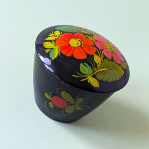 Op de afbeelding: Een kleine, ronde, zwart gelakte doos met een glanzende afwerking. Het deksel is voorzien van een handgeschilderd bloemmotief met feloranje en roze bloemen omringd door groene bladeren. Een rode bes met groene bladeren staat op de zijkant.