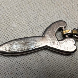 May include: Silver-toned metal pendant with a unique, abstract shape. The pendant has a textured surface and a gold-colored clasp. The text "EXCLUSIVE FORTE COMBINO" and "CONDE D'OR" are visible on the pendant.