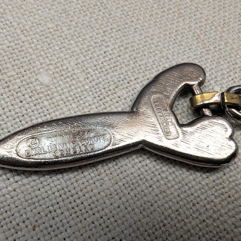 May include: Silver-toned metal pendant with a unique, abstract shape. The pendant has a textured surface and a gold-colored clasp. The text "EXCLUSIVE FORTE COMBINO" and "CONDE D'OR" are visible on the pendant.