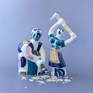 Può includere: Due statuette in ceramica con base bianca e motivi astratti dipinti a mano in blu, rosa e marrone. Una figura è in posizione seduta, mentre l'altra è in piedi con le braccia sollevate che tiene un attrezzo. Piccoli chicchi bianchi sono sparsi.
