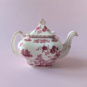 Op de afbeelding: Een crèmekleurige keramische theepot met een donkerroze bloemenpatroon in transferdruk. De theepot heeft een vierkant lichaam, een bijpassend deksel met een puntige knop, een gebogen handvat en een tuit met gedetailleerde botanische motieven.