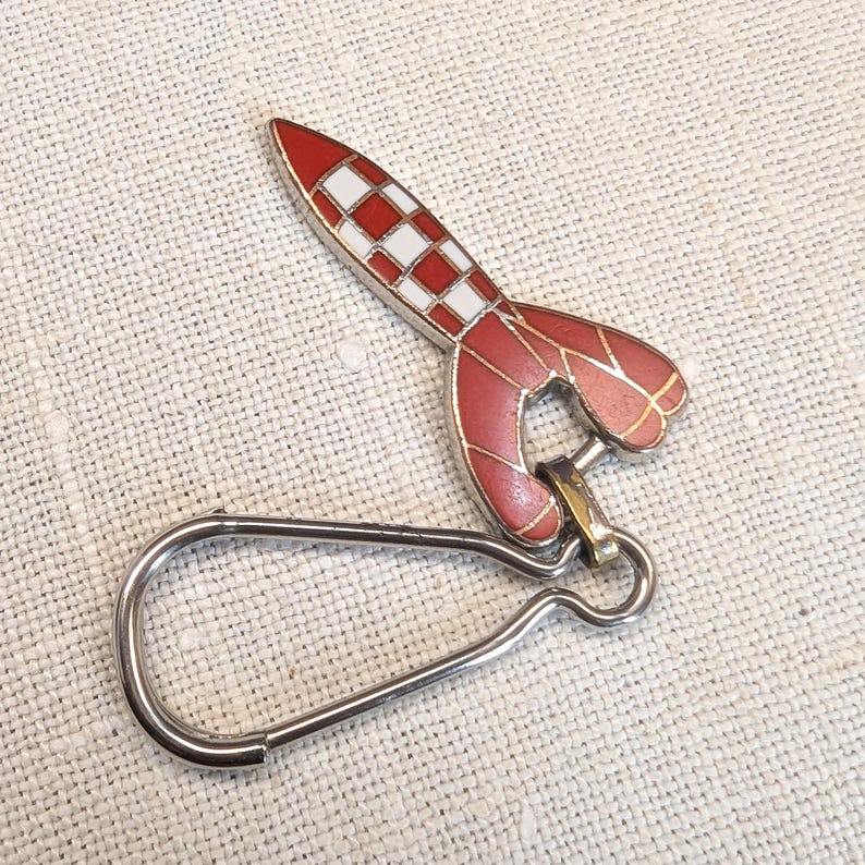 May include: A red and white rocket-shaped keychain with a silver clasp. The rocket has a checkered pattern on the top and red accents. The keychain is made of metal and is designed to attach to keys or a bag.
