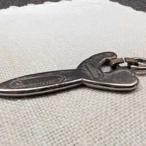 May include: A silver-colored bottle opener with a unique shape, featuring the text "USSNEL FORGE LOMBARO". The opener has a key ring attachment and a small, gold-colored metal piece. The design is detailed and the metal has a slightly aged appearance.