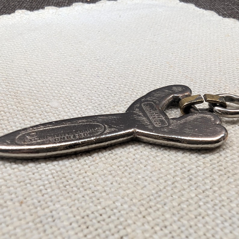 May include: A silver-colored bottle opener with a unique shape, featuring the text "USSNEL FORGE LOMBARO". The opener has a key ring attachment and a small, gold-colored metal piece. The design is detailed and the metal has a slightly aged appearance.