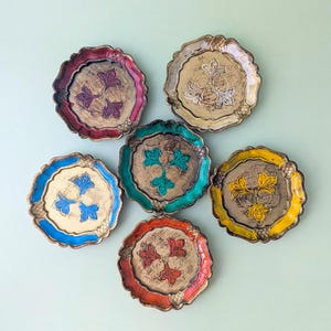 May include: Six round decorative plates with scalloped edges and a textured gold center. Each plate features a raised, three-part floral motif in a distinct color: red, white, blue, green, yellow, and orange, with matching colored rims.