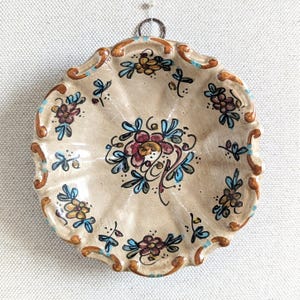 Könnte beinhalten: Ein runder, gewellter Keramik-Wandteller mit cremefarbener Glasur. Die Oberfläche zeigt handgemalte florale Motive in Blau-, Braun- und Gelbtönen, die in einer zentralen Gruppe angeordnet und entlang des Randes wiederholt sind.