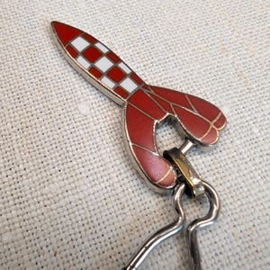 May include: A red and white rocket-shaped keychain with a silver-colored metal clasp. The rocket has a checkered pattern on the top and a stylized design on the body. The keychain is designed to be attached to keys or a bag.