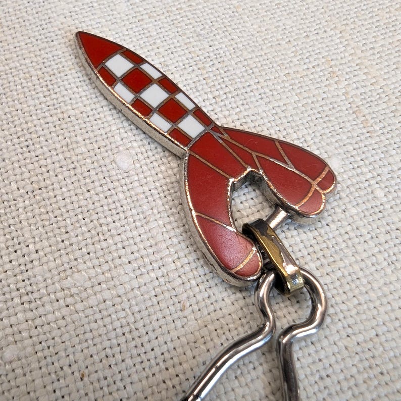 May include: A red and white rocket-shaped keychain with a silver-colored metal clasp. The rocket has a checkered pattern on the top and a stylized design on the body. The keychain is designed to be attached to keys or a bag.