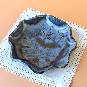 Puede incluir: Un pequeño plato de cerámica redondo con un borde ondulado y festoneado. La superficie presenta un esmalte brillante y moteado en tonos azul profundo, gris carbón y toques de bronce metálico, creando una apariencia texturizada y variada.