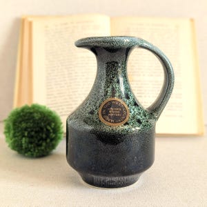 Puede incluir: Una jarra de cerámica con un esmalte moteado en azul oscuro y verde, que presenta un asa curva y una pegatina dorada circular que dice Made in the West Country Foster's Studio Pottery. La jarra tiene una base ancha y un cuello estrecho.