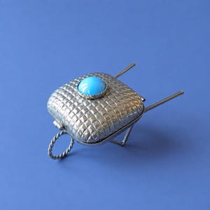 May include: A silver-toned metal miniature wheelbarrow featuring a textured, grid-patterned square container. A smooth, round, bright turquoise cabochon is set in the center of the top. The structure includes two thin handles and a twisted metal wheel.
