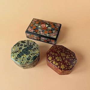 Pode incluir: Três caixas decorativas com tampas. Uma caixa retangular preta apresenta um padrão floral multicolorido vibrante. Duas caixas octogonais estão incluídas: uma com um design floral em verde escuro e dourado e outra com um padrão floral em vermelho escuro e dourado.