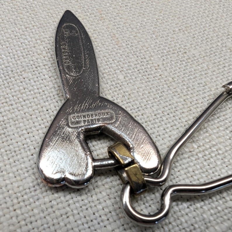 May include: A silver-toned brooch with a unique design, featuring a stylized shape with a loop and a safety pin closure. The brooch has the text "COINDEROUX PARIS" engraved on it.