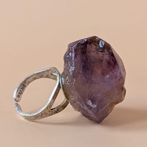 May include: A silver ring featuring a large, raw, deep purple amethyst crystal as the centrepiece. The metal band is open-ended and has the word SILVER stamped on the inner surface, showcasing a textured, metallic finish throughout the band.