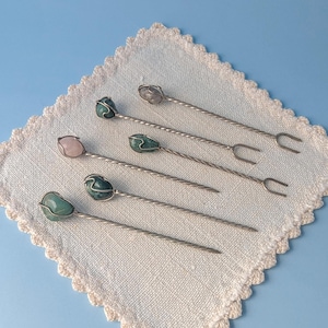 May include: Five metal hair pins featuring twisted stems and polished gemstone toppers. The stones include shades of green, a pale pink, and a translucent white, each secured with thin wire wrapping. Two pins have a two-pronged fork end.