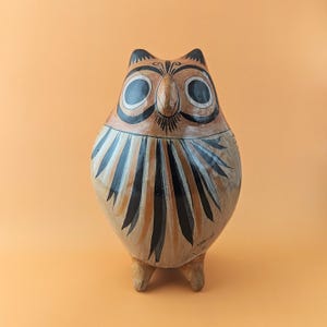 Könnte beinhalten: Eine handbemalte Eulenfigur aus Keramik mit einem abgerundeten Körper in beiger Grundfarbe. Die Eule hat große schwarz-weiße kreisförmige Augen, einen spitzen Schnabel und schwarze federartige Markierungen auf der Brust. Sie steht auf zwei kleinen Füßen.
