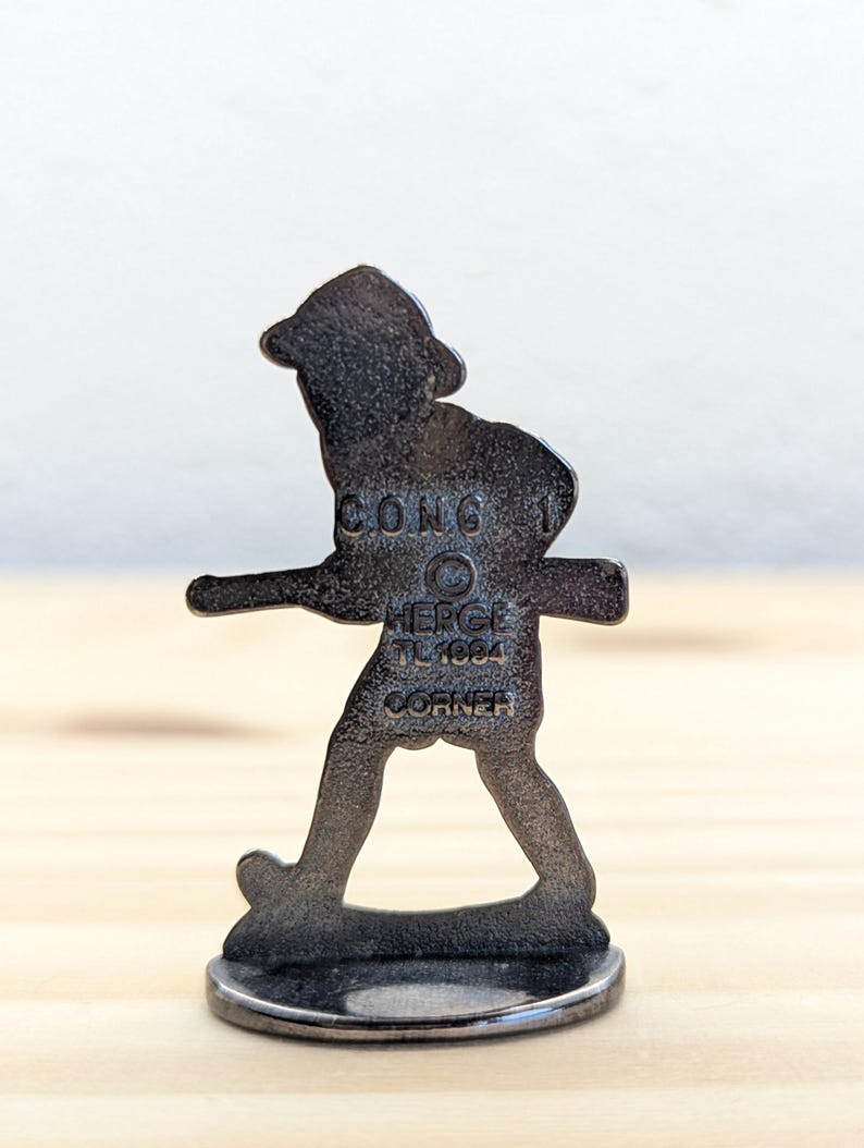 May include: A dark metal figurine of a person walking, with a hat and a stick. The figurine is on a round base. The text "CONG 1", "HERGE TL1984 CORNER" is visible on the figurine.