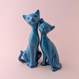 Può includere: Due statuette di gatto in ceramica blu scuro lucido, posizionate una accanto all'altra. Il gatto più alto a sinistra ha gli occhi spalancati con ciglia dipinte, mentre il gatto più piccolo a destra ha gli occhi chiusi e un'espressione serena.