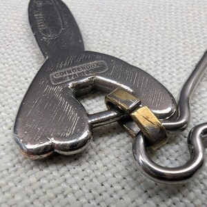 May include: Close-up of a silver-toned metal pendant with a heart-shaped design and a gold-colored accent. The pendant has the text "COINDEROUX PARIS" engraved on it. The pendant is on a white surface.