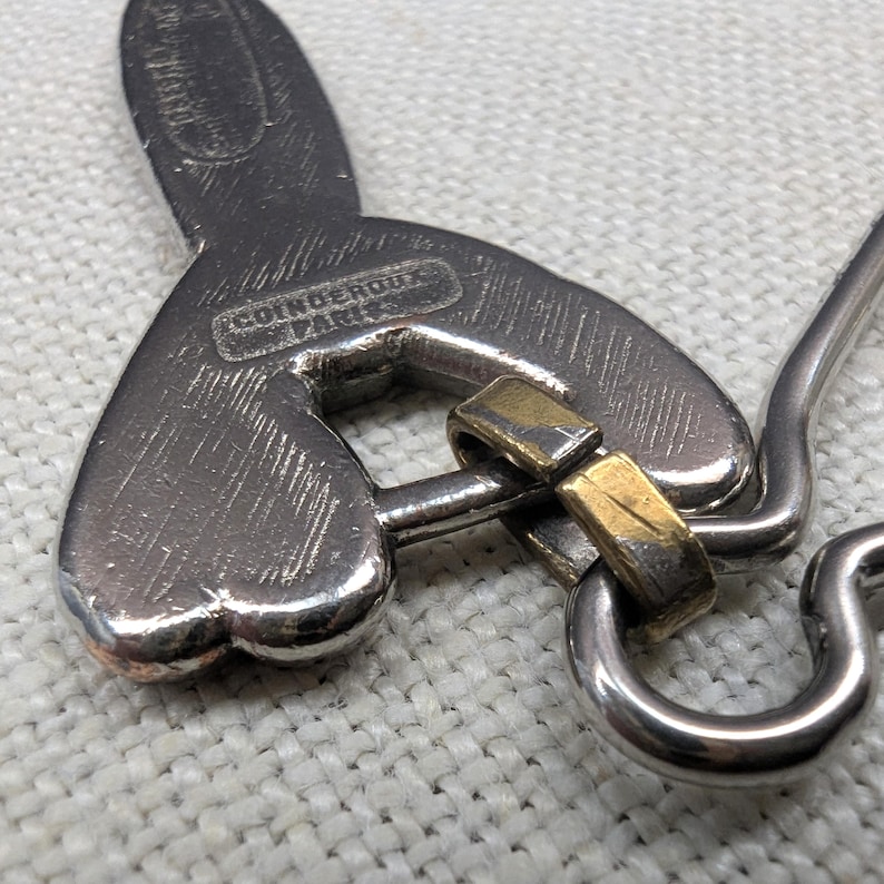 May include: Close-up of a silver-toned metal pendant with a heart-shaped design and a gold-colored accent. The pendant has the text "COINDEROUX PARIS" engraved on it. The pendant is on a white surface.