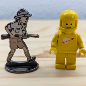 May include: Two toy figures on a wooden surface. One is a silver-coloured metal figurine of a person in a safari outfit holding a rifle. The other is a yellow Lego astronaut with a white and red logo on the chest.