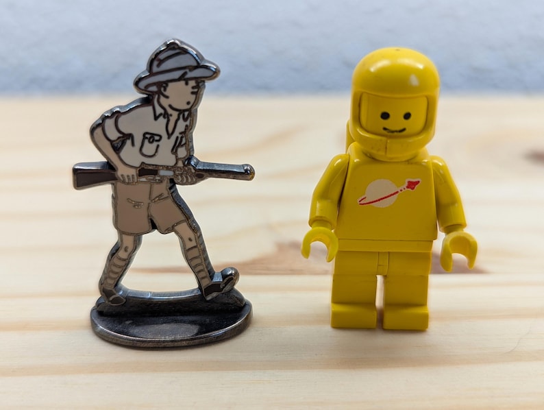 May include: Two toy figures on a wooden surface. One is a silver-coloured metal figurine of a person in a safari outfit holding a rifle. The other is a yellow Lego astronaut with a white and red logo on the chest.